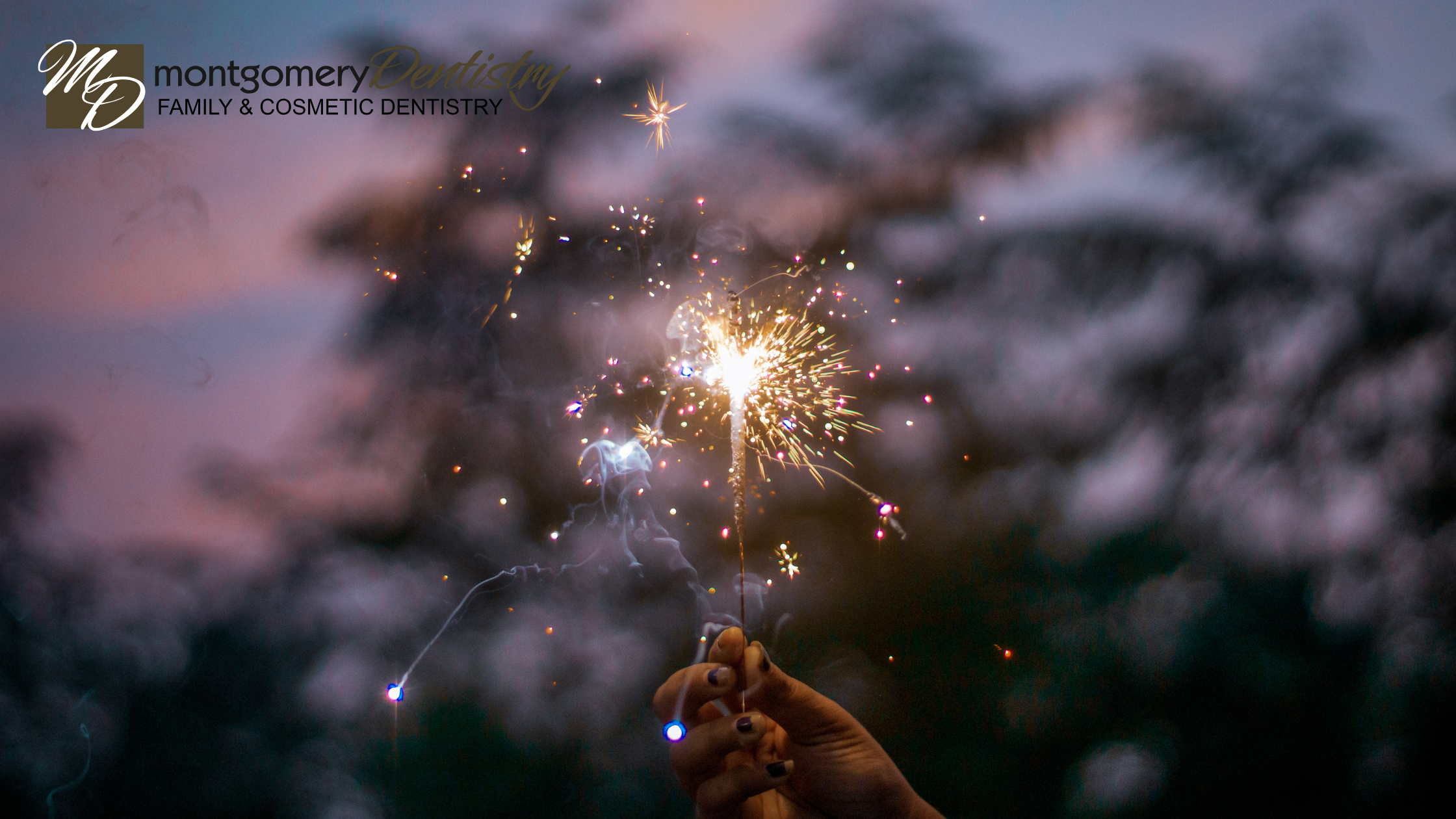 Someone's hand holding a sparkler at dusk. Trees and a pink and blue sky can be seen in the background.