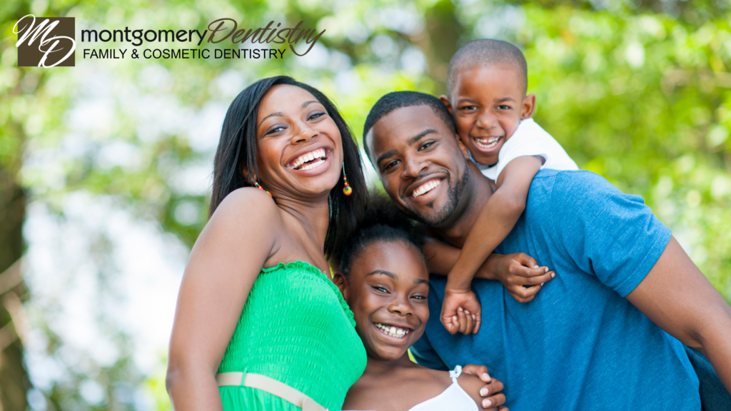 A smiling family of four—father, mother, son, and daughter—posing together in a bright, outdoor park setting. The top left corner features the Montgomery Dentistry Family and Cosmetic Dentistry logo.