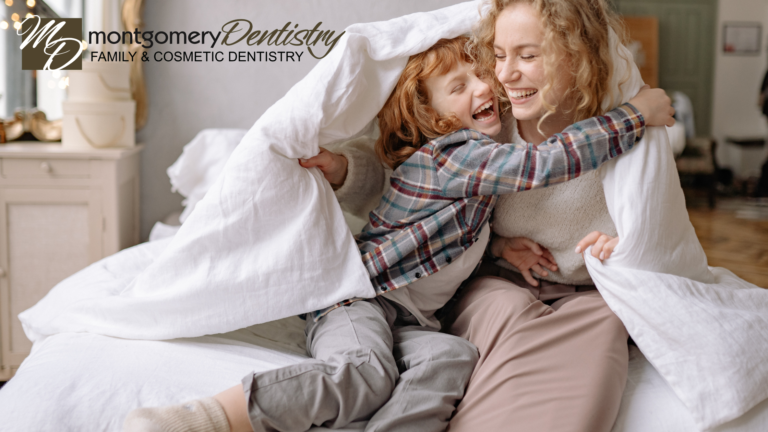 A joyful woman and young child laughing and playing together under a white blanket on a bed, capturing a happy family moment. The Montgomery Dentistry Family and Cosmetic Dentistry logo is positioned in the top left corner.